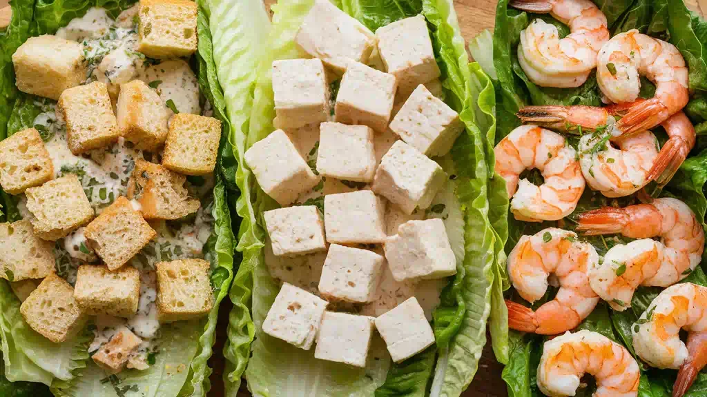 Three versions of Caesar salad: a classic with croutons, a vegan with tofu, and a hearty one with grilled shrimp arranged on wooden platters.